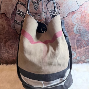 May include: A bucket bag featuring a beige canvas body with a prominent red, black, and white check pattern. The bag includes a black leather handle and a matching black leather shoulder strap, with silver-toned metal hardware attachments.