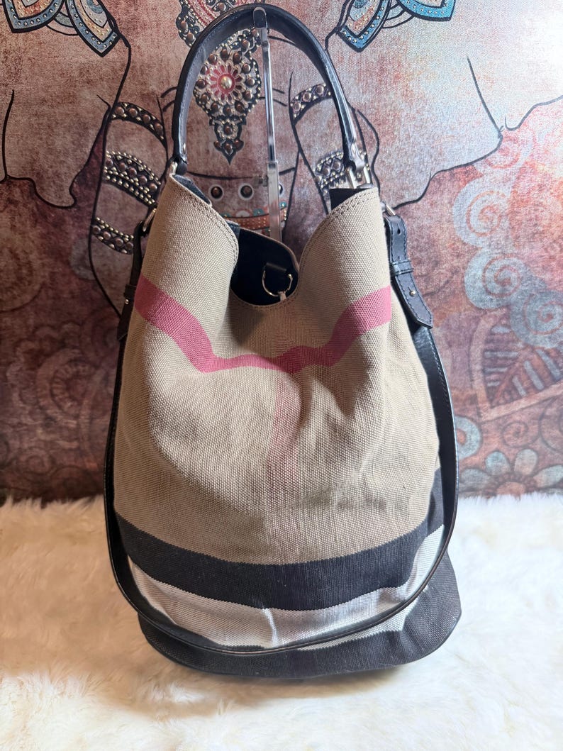 May include: A bucket bag featuring a beige canvas body with a prominent red, black, and white check pattern. The bag includes a black leather handle and a matching black leather shoulder strap, with silver-toned metal hardware attachments.