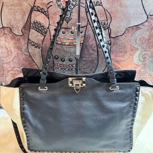 K&ouml;nnte beinhalten: Eine schwarze Ledertasche mit cremefarbenen Seitenteilen. Die Tasche verf&uuml;gt &uuml;ber silberfarbene Hardware, eine genietete Zierleiste und doppelte Schultergurte. Die Tasche hat einen oberen Verschluss und einen abnehmbaren Schultergurt.