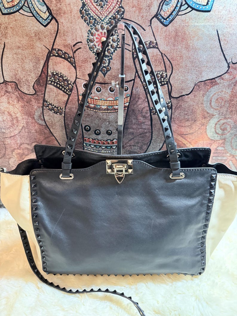 K&ouml;nnte beinhalten: Eine schwarze Ledertasche mit cremefarbenen Seitenteilen. Die Tasche verf&uuml;gt &uuml;ber silberfarbene Hardware, eine genietete Zierleiste und doppelte Schultergurte. Die Tasche hat einen oberen Verschluss und einen abnehmbaren Schultergurt.