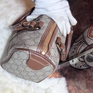 May include: A barrel-shaped handbag featuring a beige and brown monogram pattern with brown leather trim, gold-tone hardware, and a top zipper closure. The bag includes two brown leather handles attached with metal hardware and reinforced stitching.