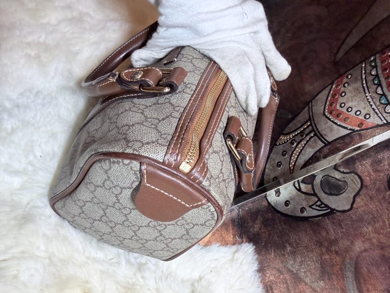 May include: A barrel-shaped handbag featuring a beige and brown monogram pattern with brown leather trim, gold-tone hardware, and a top zipper closure. The bag includes two brown leather handles attached with metal hardware and reinforced stitching.