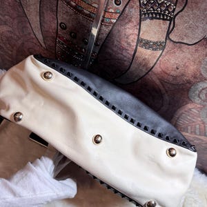 K&ouml;nnte beinhalten: Cremefarbene und schwarze Handtasche mit goldfarbenen Nieten. Die Tasche hat ein zweifarbiges Design mit einem cremefarbenen Korpus und einem schwarzen Oberteil. Sie ist mit goldfarbenen und schwarzen Pyramidennieten verziert.