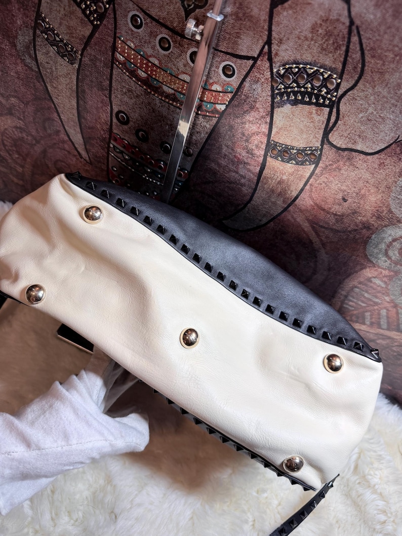 K&ouml;nnte beinhalten: Cremefarbene und schwarze Handtasche mit goldfarbenen Nieten. Die Tasche hat ein zweifarbiges Design mit einem cremefarbenen Korpus und einem schwarzen Oberteil. Sie ist mit goldfarbenen und schwarzen Pyramidennieten verziert.