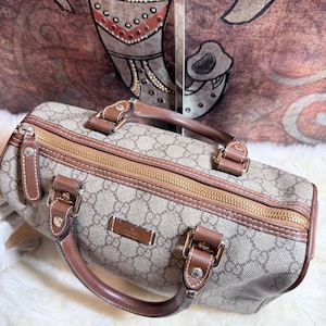 May include: A beige and brown monogram patterned handbag featuring a top zipper closure, brown leather handles, and gold-toned hardware. The bag displays a rectangular leather patch with the embossed text Gucci on the front panel.