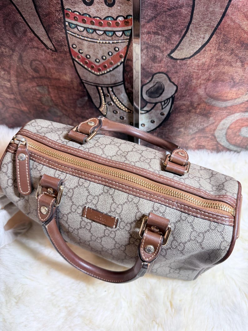 May include: A beige and brown monogram patterned handbag featuring a top zipper closure, brown leather handles, and gold-toned hardware. The bag displays a rectangular leather patch with the embossed text Gucci on the front panel.