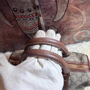May include: A beige and brown handbag featuring a repeating interlocking G logo pattern, brown leather handles, and a gold-tone zipper closure. The bag has a structured shape with brown leather trim along the edges and a metallic zipper pull.