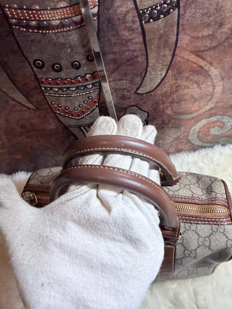 May include: A beige and brown handbag featuring a repeating interlocking G logo pattern, brown leather handles, and a gold-tone zipper closure. The bag has a structured shape with brown leather trim along the edges and a metallic zipper pull.