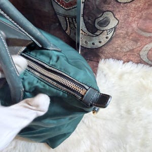 K&ouml;nnte beinhalten: T&uuml;rkisfarbene Handtasche mit silbernem Rei&szlig;verschluss und dunkelgr&uuml;nen Ledergriffen. Die Tasche hat eine runde Form und einen schwarzen Lederakzent an der Seite. Der Hintergrund ist eine wei&szlig;e, flauschige Oberfl&auml;che.