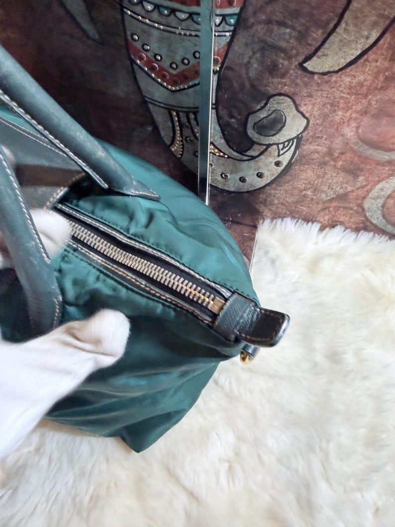 K&ouml;nnte beinhalten: T&uuml;rkisfarbene Handtasche mit silbernem Rei&szlig;verschluss und dunkelgr&uuml;nen Ledergriffen. Die Tasche hat eine runde Form und einen schwarzen Lederakzent an der Seite. Der Hintergrund ist eine wei&szlig;e, flauschige Oberfl&auml;che.
