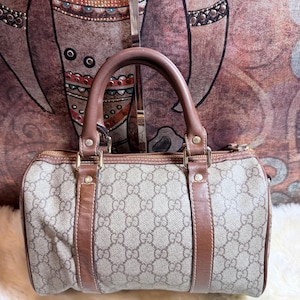 May include: A beige and brown barrel-shaped handbag featuring the iconic Gucci GG monogram pattern. The bag includes brown leather trim, two top handles, gold-tone hardware, and a top zipper closure. The exterior is constructed from a coated canvas material.