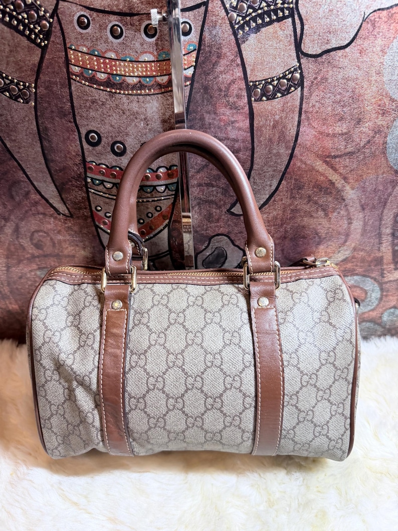 May include: A beige and brown barrel-shaped handbag featuring the iconic Gucci GG monogram pattern. The bag includes brown leather trim, two top handles, gold-tone hardware, and a top zipper closure. The exterior is constructed from a coated canvas material.