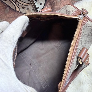 May include: A brown and beige monogram patterned handbag with a top zipper closure, brown leather trim, and a dark brown interior lining. The bag features dual leather handles attached with gold-tone hardware and a structured rectangular base.