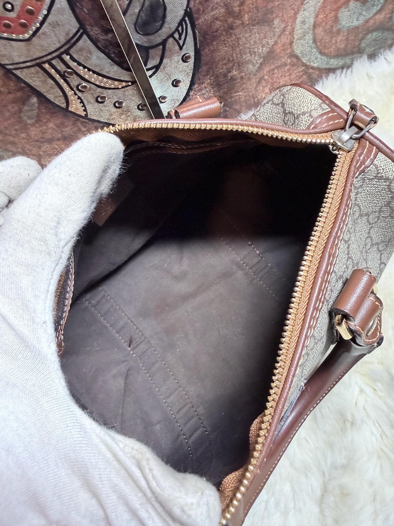 May include: A brown and beige monogram patterned handbag with a top zipper closure, brown leather trim, and a dark brown interior lining. The bag features dual leather handles attached with gold-tone hardware and a structured rectangular base.