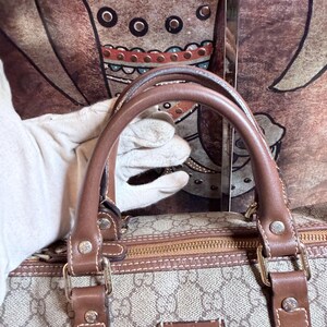 May include: A beige and brown handbag featuring the iconic GG monogram pattern, brown leather handles, and brown leather trim. The bag includes a rectangular brown leather patch with the Gucci logo embossed on the front and gold-tone hardware.