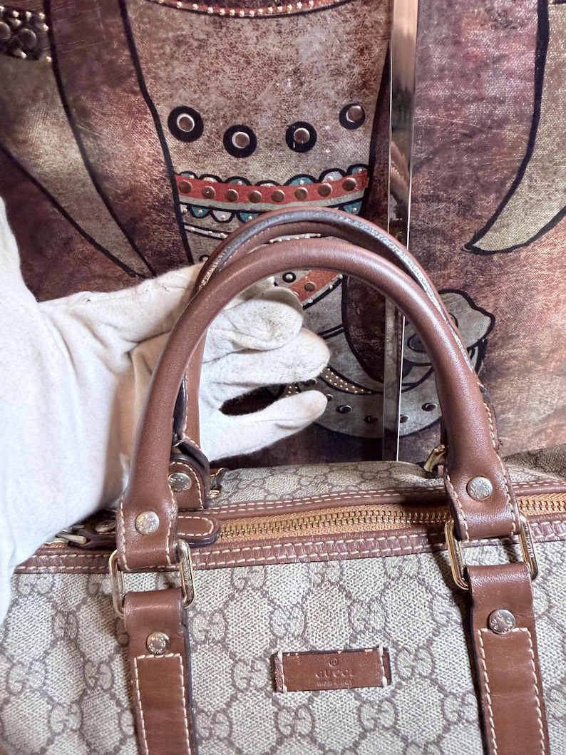 May include: A beige and brown handbag featuring the iconic GG monogram pattern, brown leather handles, and brown leather trim. The bag includes a rectangular brown leather patch with the Gucci logo embossed on the front and gold-tone hardware.