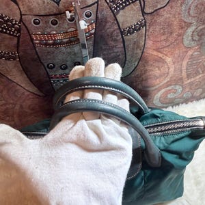 K&ouml;nnte beinhalten: T&uuml;rkisgr&uuml;ne Handtasche mit schwarzem Rei&szlig;verschluss und grauen Ledergriffen. Die Tasche besteht aus Nylon und hat einen silbernen Rei&szlig;verschluss. Die Griffe sind mit wei&szlig;em Faden gen&auml;ht. Die Tasche liegt auf einer wei&szlig;en Oberfl&auml;che.