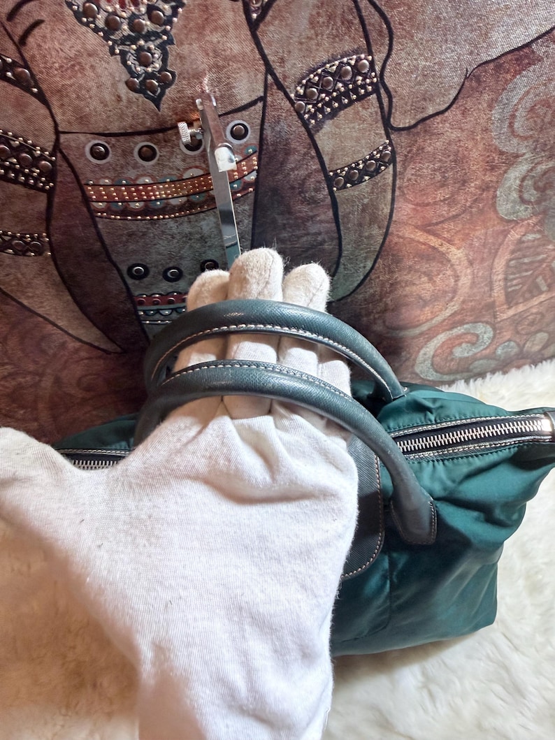 K&ouml;nnte beinhalten: T&uuml;rkisgr&uuml;ne Handtasche mit schwarzem Rei&szlig;verschluss und grauen Ledergriffen. Die Tasche besteht aus Nylon und hat einen silbernen Rei&szlig;verschluss. Die Griffe sind mit wei&szlig;em Faden gen&auml;ht. Die Tasche liegt auf einer wei&szlig;en Oberfl&auml;che.