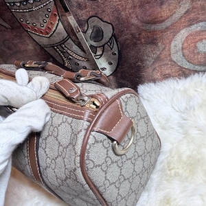 May include: A beige and brown patterned handbag featuring the iconic interlocking GG monogram print, brown leather trim, a gold-tone metal zipper closure, and a D-ring attachment on the side for a strap, showcasing a structured barrel shape.