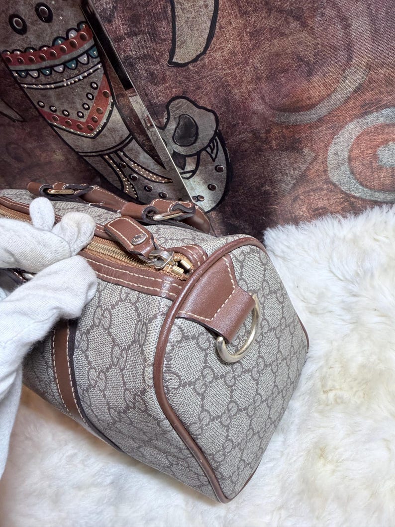 May include: A beige and brown patterned handbag featuring the iconic interlocking GG monogram print, brown leather trim, a gold-tone metal zipper closure, and a D-ring attachment on the side for a strap, showcasing a structured barrel shape.