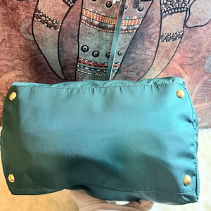 K&ouml;nnte beinhalten: T&uuml;rkisfarbene Handtasche mit goldfarbener Hardware. Die Tasche hat eine rechteckige Form mit abgerundeten Ecken und einem gesteppten Design. Die Hardware umfasst vier runde Nieten an der Unterseite. Die Tasche wird von einer behandschuhten Hand gehalten.