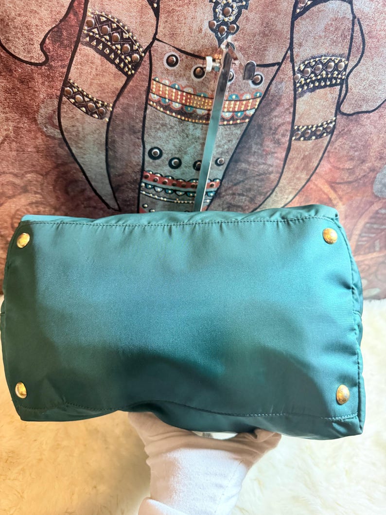 K&ouml;nnte beinhalten: T&uuml;rkisfarbene Handtasche mit goldfarbener Hardware. Die Tasche hat eine rechteckige Form mit abgerundeten Ecken und einem gesteppten Design. Die Hardware umfasst vier runde Nieten an der Unterseite. Die Tasche wird von einer behandschuhten Hand gehalten.