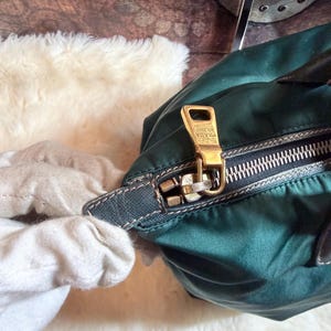 K&ouml;nnte beinhalten: Nahaufnahme einer dunkelgr&uuml;nen Prada-Handtasche. Die Tasche hat einen goldfarbenen Rei&szlig;verschluss und ein goldfarbenes Prada-Logo. Der Rei&szlig;verschluss ist ge&ouml;ffnet und gibt den Blick ins Innere frei. Die Tasche ist aus Nylon.