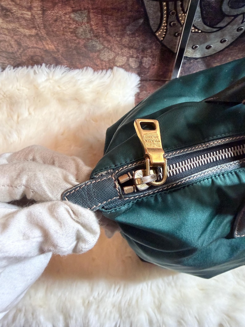 K&ouml;nnte beinhalten: Nahaufnahme einer dunkelgr&uuml;nen Prada-Handtasche. Die Tasche hat einen goldfarbenen Rei&szlig;verschluss und ein goldfarbenes Prada-Logo. Der Rei&szlig;verschluss ist ge&ouml;ffnet und gibt den Blick ins Innere frei. Die Tasche ist aus Nylon.