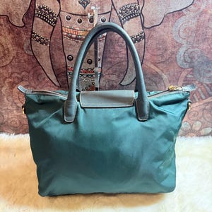 K&ouml;nnte beinhalten: Eine tealfarbene Tragetasche mit grauem Ledergriff und Klappenverschluss. Die Tasche hat einen Rei&szlig;verschluss und goldfarbene Hardware. Die Tasche ist aus einem glatten, gl&auml;nzenden Material gefertigt.