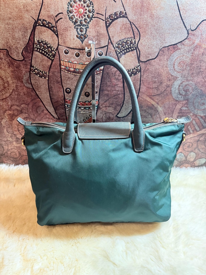 K&ouml;nnte beinhalten: Eine tealfarbene Tragetasche mit grauem Ledergriff und Klappenverschluss. Die Tasche hat einen Rei&szlig;verschluss und goldfarbene Hardware. Die Tasche ist aus einem glatten, gl&auml;nzenden Material gefertigt.