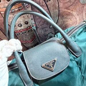 K&ouml;nnte beinhalten: Eine teal-gr&uuml;ne Prada Handtasche mit einer kleinen, grauen Klapptasche. Die Tasche hat das Prada-Logo in Gold. Die Tasche hat graue Griffe und einen Rei&szlig;verschluss. Der Hintergrund ist ein gemusterter Stoff.
