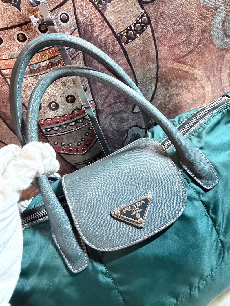 K&ouml;nnte beinhalten: Eine teal-gr&uuml;ne Prada Handtasche mit einer kleinen, grauen Klapptasche. Die Tasche hat das Prada-Logo in Gold. Die Tasche hat graue Griffe und einen Rei&szlig;verschluss. Der Hintergrund ist ein gemusterter Stoff.