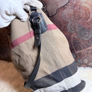 May include: A beige canvas tote bag featuring a signature plaid pattern in black, white, and red stripes. The bag includes black leather trim, silver-toned metal hardware, and a black leather shoulder strap attached with metal rings to the top.