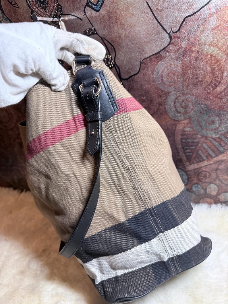 May include: A beige canvas tote bag featuring a signature plaid pattern in black, white, and red stripes. The bag includes black leather trim, silver-toned metal hardware, and a black leather shoulder strap attached with metal rings to the top.