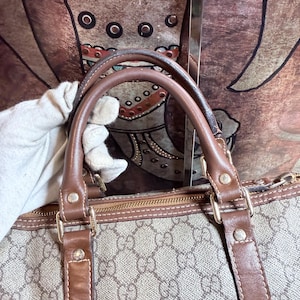 May include: A beige and brown handbag featuring a repeating interlocking G monogram pattern. The bag has brown leather handles and trim, secured with gold-tone metal hardware rivets. A zipper closure runs along the top edge of the bag.