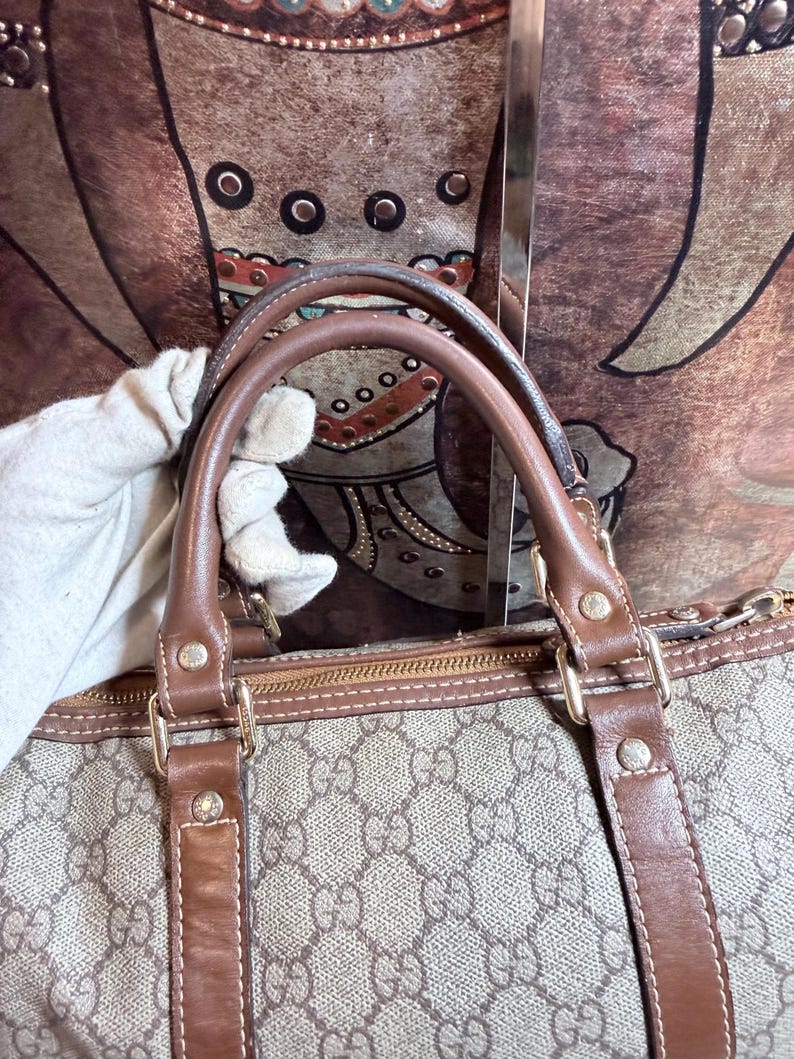 May include: A beige and brown handbag featuring a repeating interlocking G monogram pattern. The bag has brown leather handles and trim, secured with gold-tone metal hardware rivets. A zipper closure runs along the top edge of the bag.