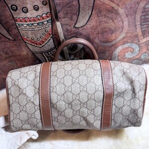 May include: A beige and brown handbag featuring the iconic GG monogram pattern across the canvas body. The bag is accented with smooth brown leather trim and two brown leather handles, showcasing a structured design with visible stitching.