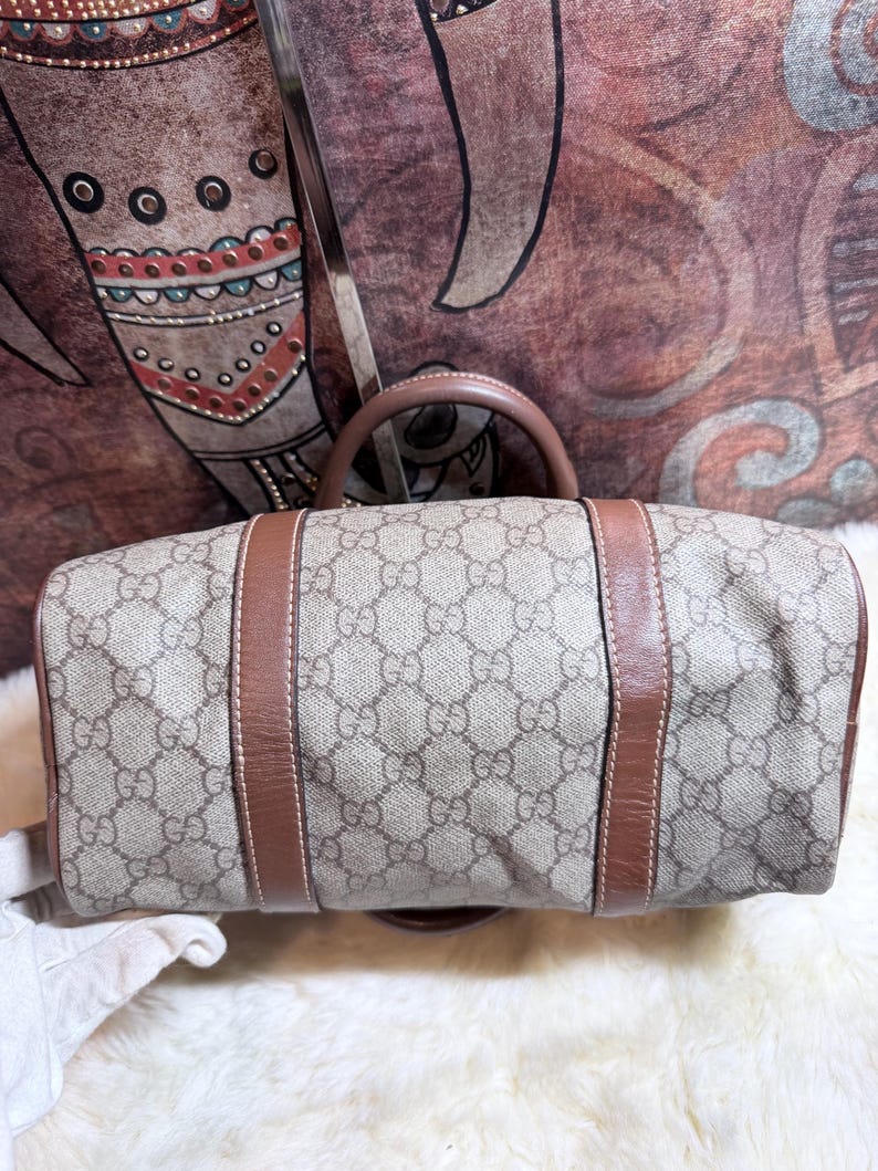 May include: A beige and brown handbag featuring the iconic GG monogram pattern across the canvas body. The bag is accented with smooth brown leather trim and two brown leather handles, showcasing a structured design with visible stitching.