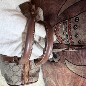 May include: A beige and brown handbag featuring the iconic GG monogram pattern, constructed with brown leather trim and sturdy rounded handles. The bag includes a gold-tone metal zipper closure along the top, showcasing detailed stitching and hardware.