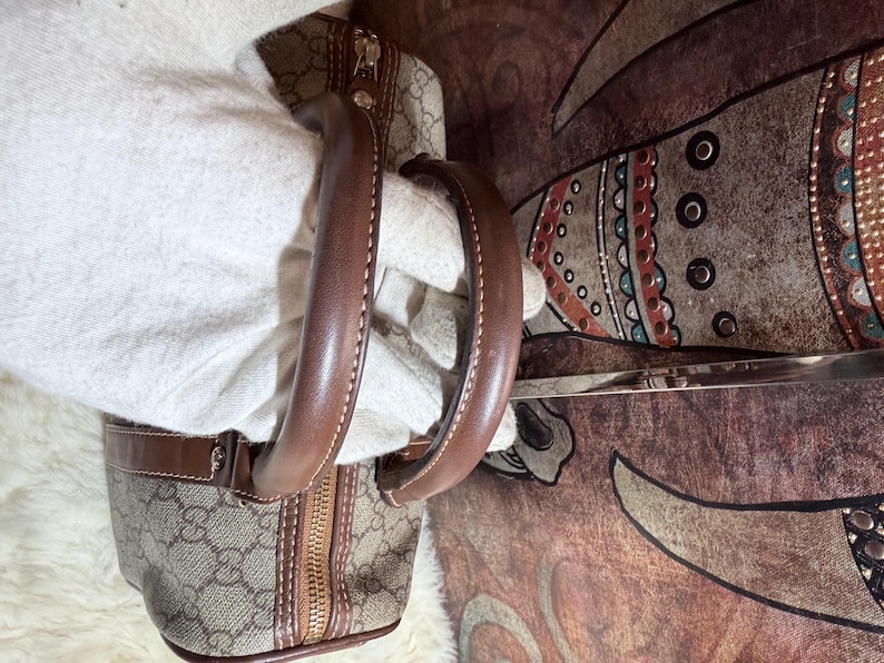 May include: A beige and brown handbag featuring the iconic GG monogram pattern, constructed with brown leather trim and sturdy rounded handles. The bag includes a gold-tone metal zipper closure along the top, showcasing detailed stitching and hardware.