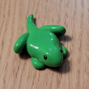May include: A small green frog figurine with black eyes. The frog is made of a smooth, glossy material and is sitting on a wooden surface.