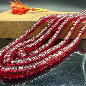May include: Strands of faceted, deep red gemstones are displayed, showcasing their reflective qualities. A tassel with red and gold threads rests on a wooden surface in the background. The gemstones are arranged in a flowing, curved pattern.