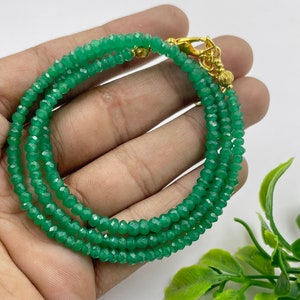 Puede incluir: Pulsera de tres hilos con pequeñas cuentas verdes facetadas. La pulsera tiene un cierre dorado. Las cuentas son de un color verde esmeralda vibrante. Se muestra sobre un fondo neutro.