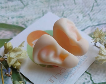 May include: Two realistic-looking, flesh-colored, sculpted human ears on a white card with the text "thank you @amarikedol" printed on it.