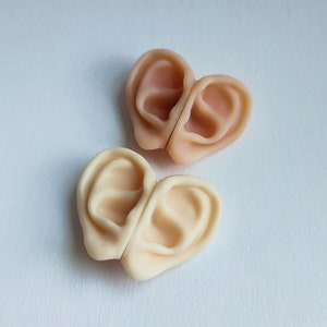 Può includere: Due paia di orecchie protesiche in una tonalità di pelle chiara, disposte a forma di cuore. Le orecchie sono dettagliate con caratteristiche realistiche, tra cui l'elica, il trago e il condotto uditivo. Lo sfondo è una superficie bianca.