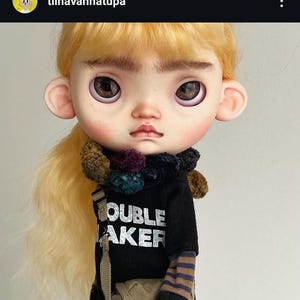 May include: A doll with blonde hair, prominent eyebrows, and large brown eyes. It wears a black shirt with the white text TROUBLE MAKER, a dark multi-colored pom-pom scarf, a brown striped long-sleeve undershirt, and a tan crossbody bag.