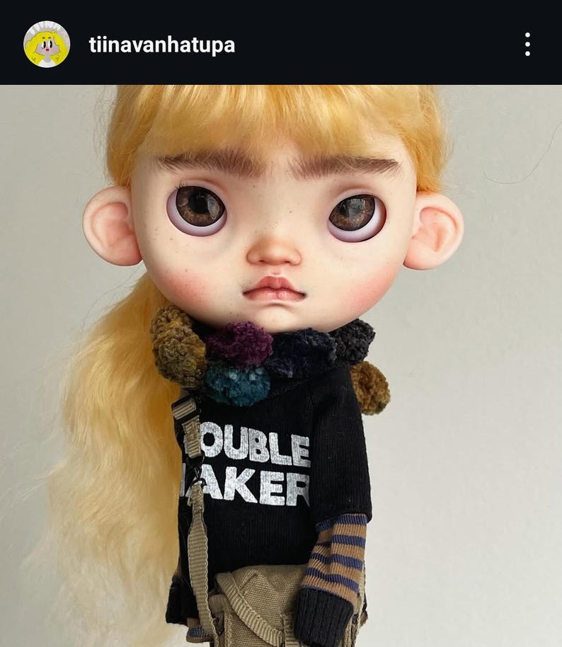 May include: A doll with blonde hair, prominent eyebrows, and large brown eyes. It wears a black shirt with the white text TROUBLE MAKER, a dark multi-colored pom-pom scarf, a brown striped long-sleeve undershirt, and a tan crossbody bag.