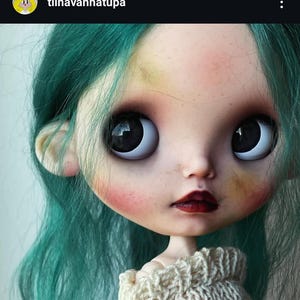 May include: A doll with teal hair, large dark eyes, and pale skin featuring freckles and rosy cheeks. The doll has dark red lips and is wearing a cream-colored knitted sweater. The text tiinavanhatupa is visible in the top left corner.