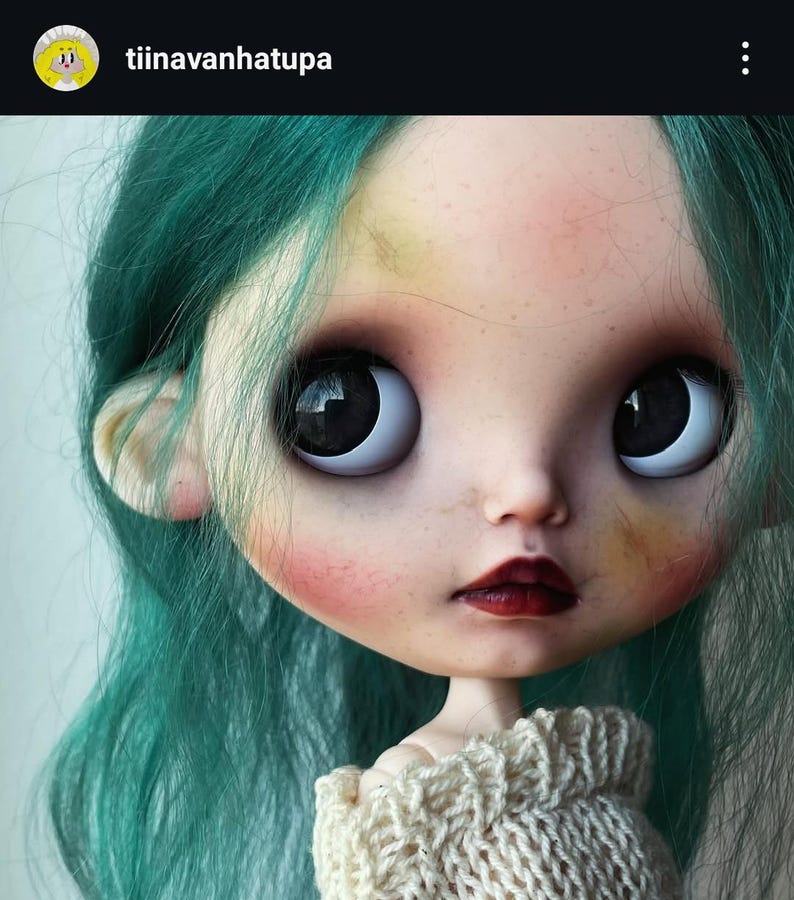 May include: A doll with teal hair, large dark eyes, and pale skin featuring freckles and rosy cheeks. The doll has dark red lips and is wearing a cream-colored knitted sweater. The text tiinavanhatupa is visible in the top left corner.