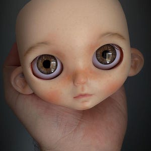 May include: A bald doll head with a smooth, pale skin tone, featuring large, round, brown eyes with detailed irises, subtle pinkish blushing on the cheeks, a small nose, and a neutral, closed-mouth expression with defined ears on the sides.